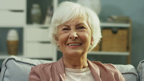 Close-up-of-the-old-lady-on-retirement-smiling-to-the-camera-while-sitting-on-the-gray-couch-in-the-living-room.-Portrait.